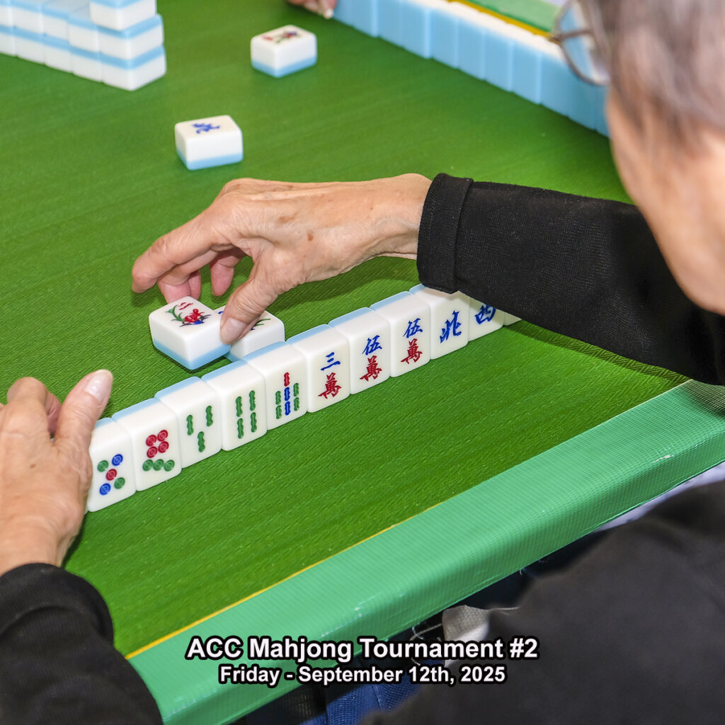 A person arranges Mahjong tiles at a green table during the ACC Mahjong Tournament #2, held on Friday, September 12th, 2025. The persons hand is reaching for a tile, and game pieces are lined up in front.