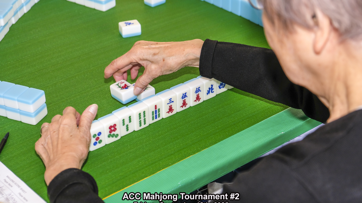 A person arranges Mahjong tiles at a green table during the ACC Mahjong Tournament #2, held on Friday, September 12th, 2025. The persons hand is reaching for a tile, and game pieces are lined up in front.