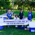 Five women sit and stand behind a Buddhist Church of Sacramento banner at an outdoor event checkpoint table, with a Checkpoint 2 sign and coolers, in a sunny park with trees in the background.