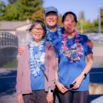 Three women wearing blue shirts and colorful leis smile and pose together outdoors at a park, with trees, a bridge, and chain-link fence visible in the background.