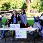 A group of six Girl Scouts stand and sit behind a table outdoors, smiling and waving. The table has signs reading “GOOD JOB!” and “girl scout troop 569.” Trees and a fence are in the background.