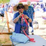 A woman wearing sunglasses and a wide-brim hat stands at a table outdoors, holding red lanyards. She is surrounded by people at an event with tents and tables under sunny weather.