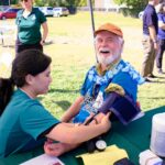 A smiling older man wearing a flower lei and baseball cap has his blood pressure checked by a healthcare worker at an outdoor event with tables, chairs, and other people in the background.