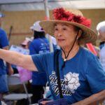 An older woman wearing a straw hat with red decorations and a blue T-shirt stands outdoors, smiling and reaching out, surrounded by other people in matching shirts at an event or festival.
