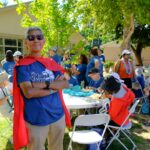 A man with gray hair and sunglasses wearing a red cape and blue Ohana Walk T-shirt stands smiling with arms crossed at an outdoor event, surrounded by people in matching shirts sitting and talking.