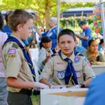 Two boys in scout uniforms stand at a table outdoors, wearing blue neckerchiefs and gloves. People are gathered in the background, some eating, under trees and tents on a sunny day.