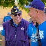 Two men smile and talk together outdoors. One man wears a military veteran cap, sunglasses, and a purple vest, while the other wears a blue shirt and cap. People and event signs are visible in the background.