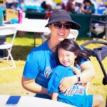 An adult and a child, both wearing blue shirts and smiling, sit together outdoors at a sunny event. The adult wears sunglasses and a black hat. White tables, chairs, and people are visible in the background.