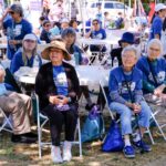 A group of older adults sit together outdoors at tables under a tent, wearing event T-shirts and hats, during a community gathering or walkathon on a sunny day.