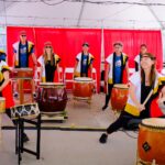 A group of people wearing traditional attire enthusiastically play taiko drums under a white canopy, with a red backdrop behind them. Several types of drums and drumsticks are visible, and the performers are smiling and energetic.