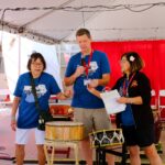 Three people stand under a tent holding microphones and papers, appearing to speak to an audience. They wear blue event t-shirts, and traditional Japanese drums are visible in the background. Red curtains decorate the space.