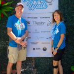 Two adults in blue Ohana Walk shirts stand smiling in front of a sponsor banner for the 2025 ACC Senior Services Ohana Walk event. The background includes greenery and a large red flower decoration.