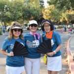Three women stand outdoors smiling, wearing matching blue Ohana Walk shirts, white skirts, visors or hats, and orange or pink bandanas. They each hold a black folding fan; trees and parked cars are in the background.