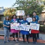 A group of seven smiling adults in matching blue shirts and colorful tutus hold handmade signs and fans, standing outdoors on a sunny day in front of a building and trees.