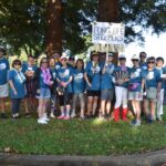 A group of people in blue Fong Bee Steppers shirts stand together outdoors, smiling, some wearing hats and sunglasses, holding a sign that reads Fong Bee Steppers, with trees and grass in the background.