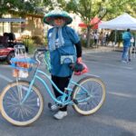 A person wearing a large blue hat, sunglasses, gloves, and a blue shirt stands in a parking lot holding a blue bicycle with a basket of oranges. Tents and people are visible in the background.