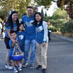 A smiling family of five, all wearing matching blue Drama Walk shirts, poses together outdoors on a sunny day. There are trees and other people in the background.