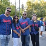 Four people wearing matching blue Nguyen 10 baseball jerseys smile and pose together outdoors. Trees and other people are visible in the background on a sunny day.