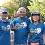Three smiling older adults wearing matching blue “Ohana Walk” shirts stand together outdoors at an event, with other participants and trees visible in the background on a sunny day.