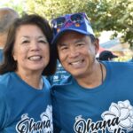 Two people wearing matching blue Ohana Walk 2023 shirts smile at the camera. The man also wears a blue hat decorated with sunglasses. There are trees and other people in similar shirts in the background.