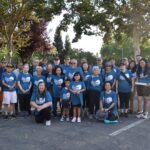 A large group of people of various ages, all wearing matching blue t-shirts, pose together outdoors in a parking lot with trees in the background on a sunny day.