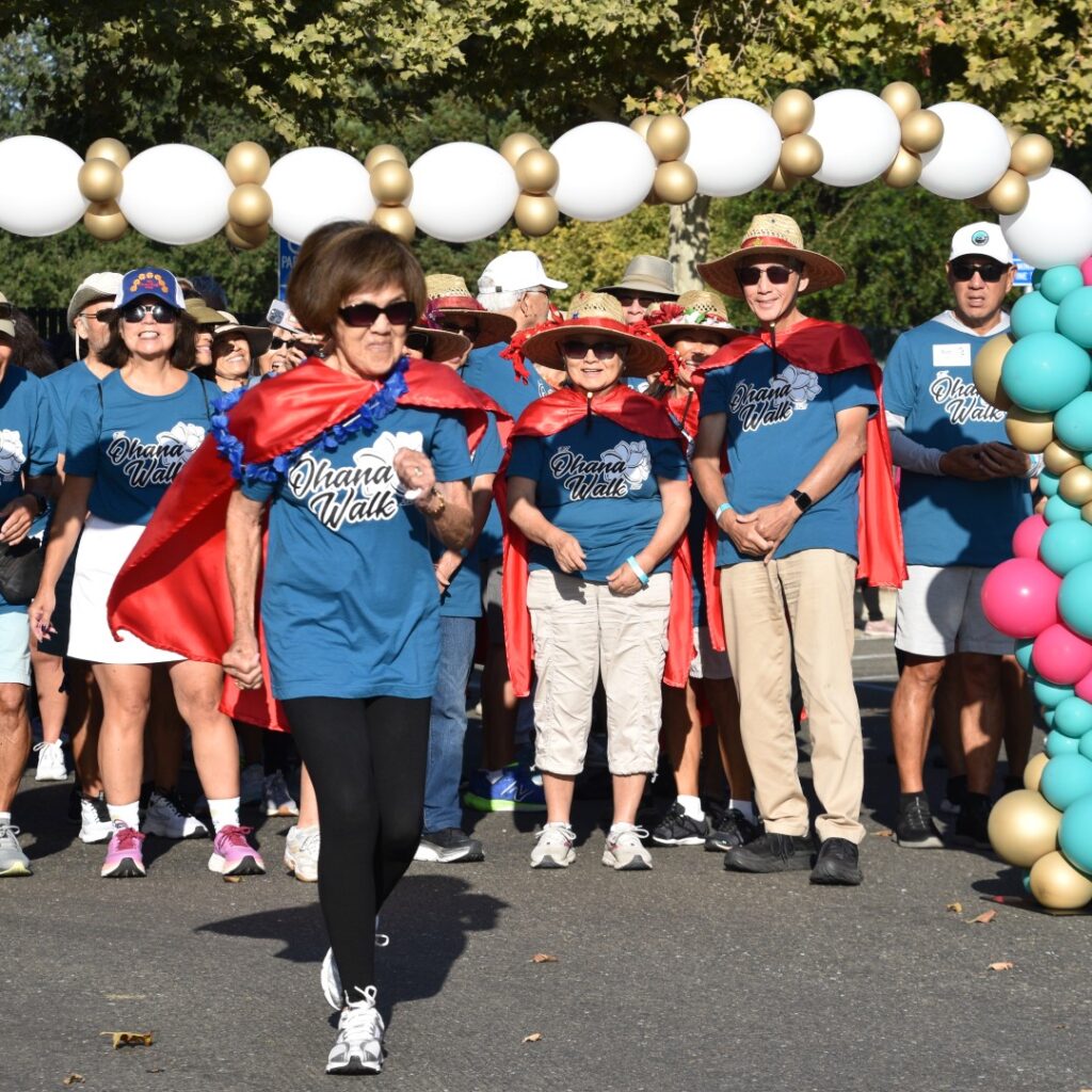A group of people wearing matching blue t-shirts and red capes stand at a starting line decorated with colorful balloons, preparing to participate in an outdoor walk or race. One woman is starting to walk ahead.