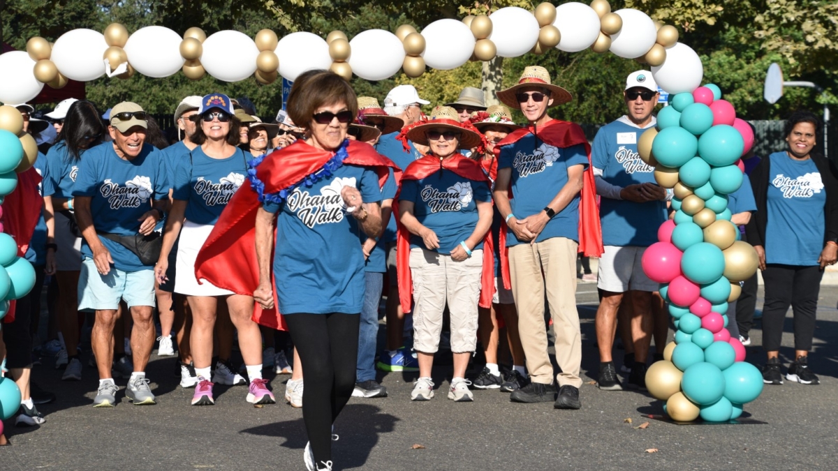 A group of people wearing matching blue t-shirts and red capes stand at a starting line decorated with colorful balloons, preparing to participate in an outdoor walk or race. One woman is starting to walk ahead.