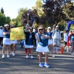 A group of people, some in blue shirts and white skirts, walk in a park event. Two women in front smile and wave; one holds a yellow sign reading Pickleballers Go for Spirit & Game! Others walk behind under trees.