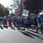 A group of people wearing blue shirts walk in a parade on a sunny street. Some hold signs, including one that says Fong’s Bee Steppers. Trees and parked cars line the road in the background.