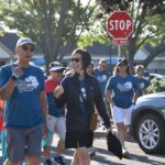 A group of people wearing matching blue Diama Walk shirts walk together outdoors on a sunny day, passing by a stop sign and parked cars, with trees and buildings in the background.