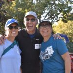 Three adults stand outdoors, smiling with their arms around each other. They wear sunglasses, casual clothes, and name tags. Other people and trees are visible in the sunny background.