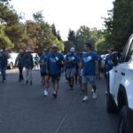 A group of people wearing blue shirts walk together on a tree-lined street during a community event, with parked white cars visible on the right side of the image.