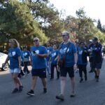 A group of people wearing matching blue shirts walk together on a sunny day along a street lined with trees. Some are wearing sunglasses, and others carry items as they participate in what appears to be an organized event.