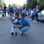 A woman kneels and hugs a small child holding a stuffed animal at an outdoor walk event, surrounded by a diverse group of people walking on a tree-lined street. Many participants wear matching blue shirts.