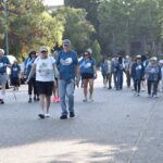 A group of people, some wearing matching blue shirts, walk together down a street lined with trees and parked cars during daylight. Some participants use canes or wear hats and sunglasses.