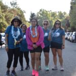 A group of five women wearing event t-shirts and leis smile for the camera while standing on a street; several other participants and parked cars are visible in the background.