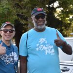 Two smiling men wearing sunglasses and Ohana Walk 2024 t-shirts stand outdoors, giving thumbs up. Trees and parked cars are visible in the background.