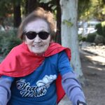 An elderly woman wearing sunglasses, a red cape, and a blue T-shirt smiles while outdoors on a sunny day, with trees and a path in the background.