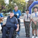 A woman holding a small dog, a man in a wheelchair, an older man standing behind him, and two teenage boys in Boy Scout uniforms pose together outdoors at an event. Trees and an inflatable arch are in the background.