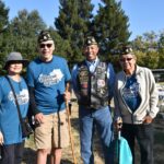 Four smiling adults stand together outdoors, wearing blue Vagana Walks shirts and veterans hats. One holds a cane, another wears a vest with patches. Folding chairs and trees are visible in the background.