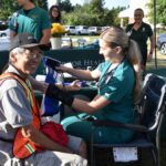 A nurse in green scrubs takes a seated older mans blood pressure at an outdoor health fair. The man smiles at the camera, wearing a hat and safety vest. A table with flowers and health staff is visible in the background.