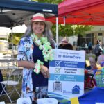 A smiling woman in a hat and floral lei stands at an outdoor booth with a sign advertising in-home care services. The booth has colorful decorations and people are gathered in the background under tents.