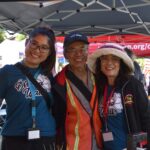 Three smiling adults pose together at an outdoor event booth. Two women wear matching blue shirts and badges, while the man in the middle wears a cap, glasses, and an orange safety vest. Tents and people are visible in the background.