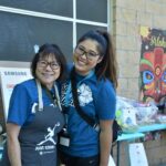 Two women smiling and posing together at an event. Both wear blue shirts, glasses, and name tags; one wears an apron. Gift baskets and an Aloha sign with a colorful tiki mask are displayed on a table behind them.