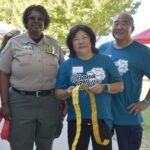Three adults smile outdoors; one wears a park ranger uniform, and the other two wear matching blue Ohana Walk shirts. The woman in the center holds a strip of yellow raffle tickets. Tents and trees are visible in the background.
