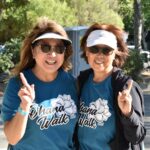 Two women wearing sunglasses and visors smile at the camera, each holding up one finger. They wear matching blue Ohana Walk 2023 T-shirts and stand outdoors with trees and sunlight in the background.