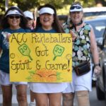 Four smiling women walk outside, with the front two holding a yellow sign that reads ACC Pickleballers Got Spirit & Game! decorated with pickleball paddles and flowers. The women wear sporty outfits and visors.