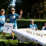 Four older adults at an outdoor event table marked Checkpoint 7 in a park; two stand and smile, one waves, and two sit behind the table with water bottles on display. All wear blue event shirts.