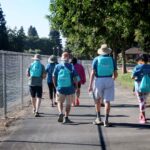 A group of people wearing matching blue shirts and backpacks walk along a paved path beside a chain-link fence and trees on a sunny day.