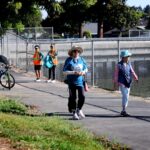 People walk along a paved path next to a chain-link fence on a sunny day. Some wear hats and vests, and a few are sitting near bicycles under the shade of trees.